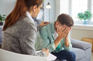 Psikolog randevusu ve terapist ile depresyondaki adamın konsültasyonu. Üzgün hasta kanepede oturuyor, elleriyle yüzünü kapatıyor, kadından destek alıyor ve rahatlatıcı dokunuşlar alıyor.