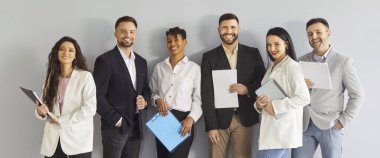 Bir grup mutlu iş adamı arkadaş canlısı iş arkadaşları, resmi kıyafetler giyiyorlar, beyaz duvara karşı poz veriyorlar, pano tutuyorlar, takım olarak çalışıyorlar, iş yerinde birlik desteğini gösteriyorlar.