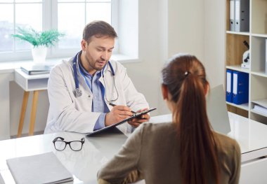 Tıbbi muayene sırasında klinikteki masada oturan hasta şikayetlerini dinleyen ve yazan doktor portresi. Hasta kadın bir doktoru ziyaret ediyor. Sağlık ve Tıp Konsepti.