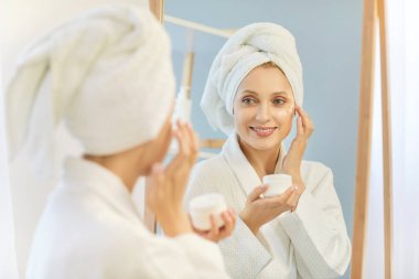 Mutlu sarışın kadın evde cilt bakımı için kavanozdan krem sürüyor. Yumuşak bornoz ve havlu giymiş bir kız aynanın karşısında duruyor ve yansımaya gülümsüyor, yüzüne masaj yapıyor.