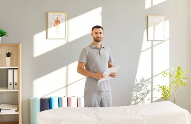 Rehabilitasyon kliniğinde kendine güvenen genç bir fizyoterapistin portresi. Erkek profesyonel fizyoterapist rehabilitasyon hastanesinde ve kamera karşısında neşeli görünüyor. Fizyoterapi kavramı