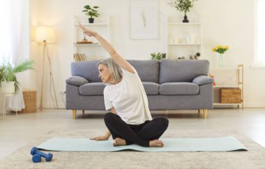 Son sınıf öğrencisi kadın evde esneyerek spor antrenmanına hazırlanıyor. Isınma rutini, esneklik ve sağlığın önemini vurgulayarak, spor ve eğitime olan bağlılığı yansıtır..