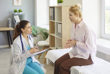 Doktor, bir klinikteki muayene sırasında hastanın semptomlarını anlatırken onu dinler, bir not panosuna not alır ve sağlık hizmeti tavsiyesi verir. Sakin tartışma doğru hasta bakımını sağlar.