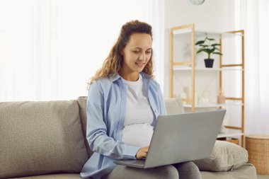 Hamile kadın oturma odasında rahat bir koltukta oturuyor, dizüstü bilgisayarla çalışıyor, ebeveynlik bilgilerini okuyor, bebek kıyafetleri sipariş ediyor, sanal teletıp hamilelik danışmanlığı yapıyor.
