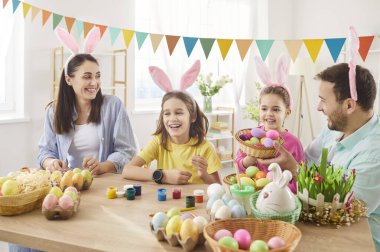 Mutlu aile, Paskalya 'yı evde birlikte kutlamak için masanın etrafında toplandı. Neşeli gülümseyen ebeveynler ve çocuklar tavşan kulağı takıyor, renkli yumurtaları parlak festival odasında dekore ederek eğleniyorlar..