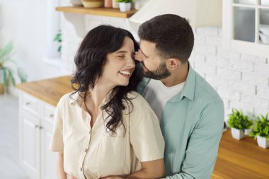 Sevgi dolu bir çift, parlak bir ev mutfağında kucaklaşır. Gülümsemeleri ve yakınlıkları, doğal ışık ve romantizmle dolu sakin bir sabahın tadını çıkarırlarken mutluluğu yansıtıyor. Sıcak aşk ve gülümseme..