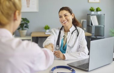 Gülümseyen doktor klinikte el sıkışan hastayı karşılıyor, randevusu sırasında steteskop ve dizüstü bilgisayarla sağlık danışmanlığına başlıyor. Sıcak karşılama güven ve yakınlık oluşturur.