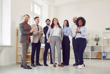 Farklı etnik çeşitlilikte iş yapan bir grup erkek ve kadın kameraya bakıp ofiste alkışlıyor ve gülümsüyor. Şirket çalışanları ya da bir grup personel iyi bir anlaşma yapmaktan mutluluk duyar..