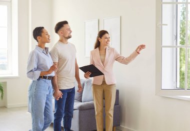 Emlakçının aile üyelerine boş bir daire gösterirken, pencereden güzel bir manzara gösterirken, modern geniş oturma odasında dururken çekilmiş bir fotoğraf. Emlak ve mülkiyet kavramı