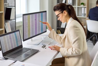 İş muhasebecisi bilgisayar analizi ile denetleme yapar. Ofisteki çift monitörlerin verilerini inceliyor, hesap tablosu verilerini kontrol ediyor ve raporlamayı doğruluyor. Mali doğruluk kavramı.