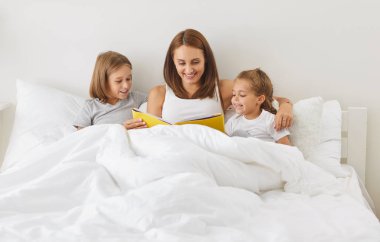 Happy family reading lying in bed, happy mother parent, cute small kids laughing together, children bonding with mother having fun enjoy funny moments in bedroom, mum telling funny fairy tale story
