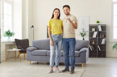 Mutlu gülümseyen genç çift ev anahtarlarını tutarak oturma odasında kameraya bakarak taşınma günü kutlamalarının tadını çıkarıyor. Taşınma, ipotek konsepti.