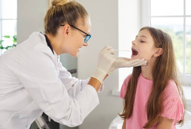 Çocuk doktoru, klinik kontrolü sırasında çocukların boğazını muayene eder. Doktor otolaringolog bademcikleri incelemek ve klinikteki enfeksiyon riskini değerlendirmek için steril bez kullanır. Pediatrik muayene.
