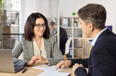 Ajan ve müvekkil ofiste sözleşmeyi tartışıyor. Gülümseyen yönetici ve danışman evrakları gözden geçirir ve modern bir masada dizüstü bilgisayarlı şartları özetlerler. Danışmanlık ve toplantı açık bir anlaşmayı güçlendirir.