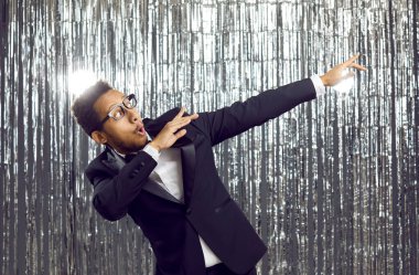 Genç ve mutlu bir etnik erkek, müziğe taşınıyor ve partide eğleniyor. Şık takım elbiseli, gömlekli ve papyonlu komik enerjik melez adam gümüş folyo fringe stüdyosunun arka planında dans ediyor.