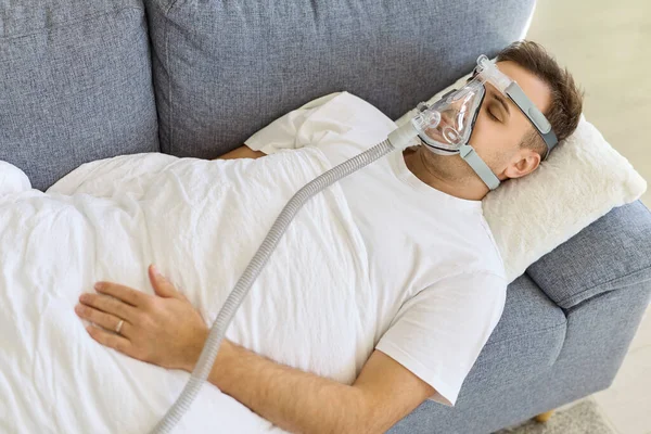 Aspiratörlü, oksijen maskeli bir adam yatakta dinleniyor. Hasta tedavi sırasında solunum desteği alıyor. Temiz kurulum, evde hastane sağlık hizmetleri olduğunu gösteriyor. Sakin tedavi konsepti.