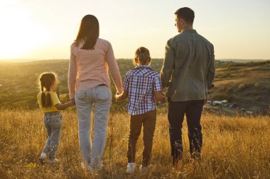 Gün batımında mutlu bir aile, dışarıda çocuklara sarılıyor. Altın ışık, doğa onları sardıkça çayırları ısıtır, tepelere tepeden bakar, sakin bir anda gülümsemelerini paylaşır. Aşk ve mutluluk kavramı.