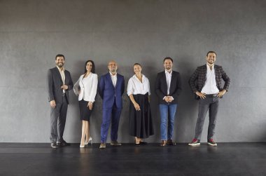 Gülümseyen iş adamları, liderliği ve takım çalışmasını vurgulayan bir şirket portresi için beton bir duvara karşı dururlar. Altı profesyonel birleşmiş, kendinden emin ve çeşitli.