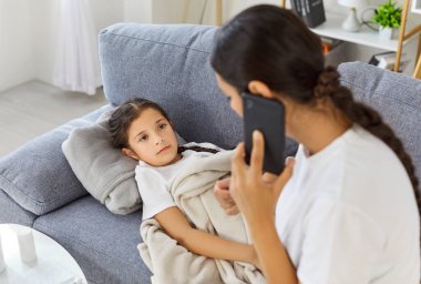 Evde kanepede yatan hasta çocuk, soğuk algınlığı ve grip tedavisi için tavsiye isteyen bir anne. Ebeveyn, kızına bakarken çocuk doktoruyla ateş hakkında telefonda konuşuyor.