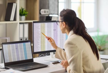 Muhasebeci ofis bilgisayarında denetim analizi yapar. Kadın verileri gözden geçirir, hesap makinesi kullanır, modern ofiste doğruluğu kontrol eder. Açık konsept finans analitik doğruluğu.