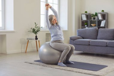 Kıdemli sporcu yogi kadın fitness topunda oturuyor oturma odasındaki yoga minderinde egzersiz yapıyor fiziksel aktivite ve esneklik, rahat ev zindeliği ve refah, sakin sağlıklı egzersiz.