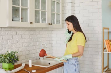 Genç bir kadın mutfakta lavabo pompasını tutuyor ve lavabonun tıkanmasını açmaya çalışan bir tesisatçıyla cep telefonuyla konuşuyor. Evdeki lavaboyu temizleyen kız. Oda servisi ve ev işleri konsepti.