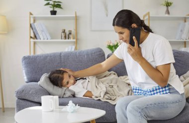 Soğuk algınlığı ve grip belirtileri olan hasta bir çocuk için doktorla telefon görüşmesi. Annesi çocukların ateşini ve ateşini ölçüyor. Aile, virüslü ev hakkında tavsiye almak için hastaneyi arıyor. Kızlarının başını tutuyor.