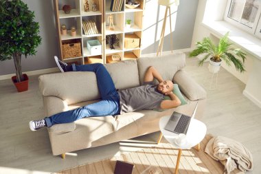 Evdeki rahat bej koltukta uyuyan rahat adam. Adam işine ara verip rahat modern oturma odasının yanındaki kahve masasında dizüstü bilgisayarla koltukta kestiriyor. Yüksek açı. Dinlenme kavramı