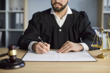 Yargıç, ahşap masada tokmak, kanun ve adalet terazileriyle yasal belgeleri imzalar. Yargıcın duruşma davasını ve mahkeme kararını, iş anlaşmasını onaylamak için profesyonel evrakları