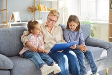 Sevgi dolu bir büyükanne öğretmenlik yapıyor, torun açık bir kitap tutuyor kanepeye oturuyor, büyükanne, aile bakıcısı ve küçük kız, erkek çocuk tatlı çocuklarla vakit geçiriyor, anaokulu torunu.