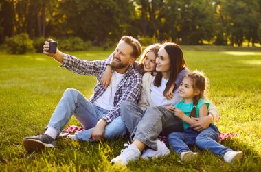 İki çocuklu gülümseyen ebeveynler güneşli bir parkta çimlerin üzerinde oturup, birbirlerine sarılıp selfie çekiyorlar. Dört kişilik mutlu aile dışarıda eğleniyor, neşeli anları kamerayla yakalıyorlar..