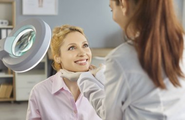 Kadın kozmetik uzmanı, güzellik salonu çalışanı 30 'lu yaşlardaki gülümseyen kadın müşterinin yüzünü inceliyor, güzellik prosedüründen önce ya da sonra cilt durumunu kontrol ediyor.