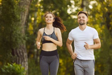 Sportif fit erkek ve kadın güneşli parkta birlikte koşuyor gülümsüyor, yeşil ağaçlar, egzersiz yaparken mutluluk, açık havada sağlıklı aktif yaşam tarzı, aile bağları ve canlılık.