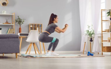 Evde spor salonunda, spor yaparken spor yaparken spor aletleri ve direnç bandı kullanan mutlu bir kadın. Aktif ve pozitiftir, spor yapmaya ve fiziksel güç eğitimine bağlılığını gösterir..