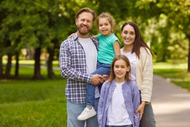 Mutlu gülümseyen aile portresi. Oğlu ve kızı yaz parkında dikilip dışarıdaki kameralara neşeli bir şekilde bakıyorlar. Genç anne, iki çocuklu baba doğa yürüyüşü yapıyor.