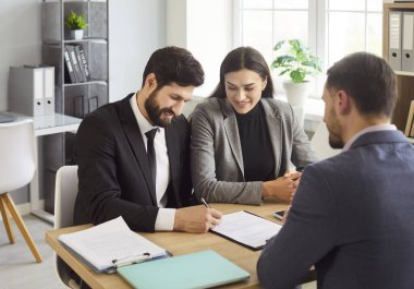 Masada oturan ve modern ofiste erkek müdürle sözleşme imzalayan mutlu genç çift. Gülümseyen adam ve kadın karar veriyor, emlakçı ya da finans danışmanıyla anlaşmaya varıyorlar..