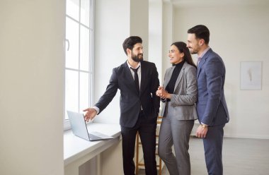 Neşeli emlakçı dizüstü bilgisayarın yanında duruyor ve pencerenin yanında gülümseyen genç çifte sunum yapıyor. Erkek emlakçıyla emlak almak ya da kiralamak için emlak seçeneklerini araştıran mutlu erkek ve kadın..
