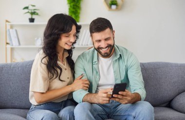 Mutlu çift evde akıllı telefon kullanıyor. Oturma odasındaki kanepede oturan gülümseyen adam ve kadın, çevrimiçi alışveriş siparişi vermek ve satın almak için ödeme yapmak, arkadaşlara video çağrısı