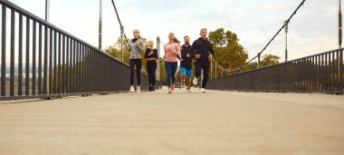 Koşucu grubu dışarıda köprüde koşuyor. Düşük açı, şehir parkında sabit tempo ve ritmi gösteriyor. Olay yerinde takım çalışması ve fitness eğitimi veriliyor. Takım çalışması ve sağlık kavramı gösterildi.