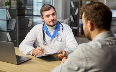 Klinikteki doktor ve hasta danışmanlığı. Steteskoplu bir doktor, not defterine not yazıp dinleyerek rutin kontrol sırasında empati ile tavsiye veriyor. Sağlık hizmetleri güven ve destek kavramı.