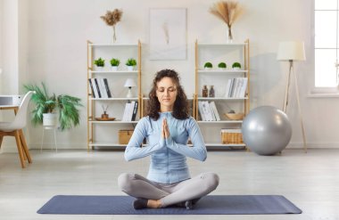 Sakin, odaklanmış bir kadın yoga ve meditasyon yapıyor, Anjali Prayer Mudra yapıyor ve Sukhasana 'da bacak bacak üstüne atarak oturuyor oturma odasının iç kısmında poz veriyor, sağlığını ve sağlıklı yaşam tarzını destekliyor.