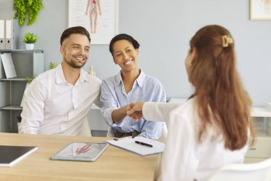 Doktor, gülümseyen çift hastayla el sıkışıyor, başarılı tıbbi konsültasyonlar, olumlu ilişkiler, karşılıklı güven ve memnuniyet, modern klinikte destekleyici bir sağlık ortamı yaratıyor.