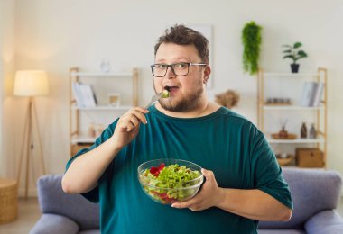Evde çatalla salata yiyen bir adam. Büyük boy gözlüklü gülümseyen yetişkin rahat bir odada bir kase marul ve domates tutuyor ve dengeyi kucaklıyor. Konsept: dikkatli beslenme ve olumlu alışkanlıklar.
