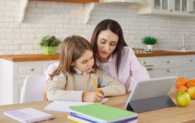 Anne, öğretmen ve öğrenci kızı internetten tablet bilgisayar kullanarak, masada birlikte oturarak, ev ödevlerine odaklanmış bir şekilde öğrenim görüyorlar.