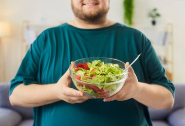 Gülümseyen adam evde salata kasesi tutuyor. Yakın plan temiz bir kasede taze marul ve domatesle el ele tutuşup sağlıklı beslenme alışkanlıklarını ön plana çıkarıyor. Kavram: sağlıklı beslenme ve günlük sağlık.