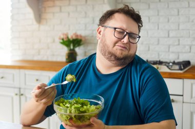 Mutfakta sebze salatası yemekten kaçınan üzgün şişman bir adam. Sağlıklı beslenme konusunda komik suratları olan tombul insan çataldan salatalık dilimleriyle uzaklaşıyor, kilo verme seçimleriyle boğuşuyor.