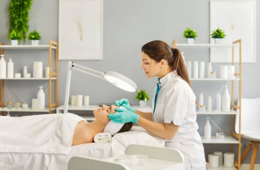 Kozmetik uzmanının hastası cilt muayenesi için randevu, profesyonel güzellik tedavisi. Yalancı kadının yüzünde el ele tutuşan bir güzellik uzmanı büyüteçten bakıyor. Kozmetoloji kavramı