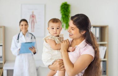 Annesi klinik kontrolünden sonra bebeği kucağında tutuyor. Gülümseyen anne, çocuk doktorunun arkasında durup iyi bir ziyaret ve aile bakımını önererek çocuğu kucağına alır. Güven kavramı, pediatrik sağlık ve tıp..