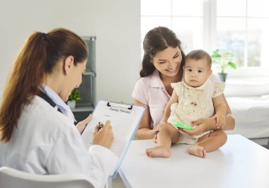 Çocuk doktoru danışmak için anne ve bebekle tanışır. Çocuk hasta klinikte oyuncaklarla oynarken aile doktoru panodaki terapi listesini inceliyor. Pediyatrik güven, bakım ve ilaç.