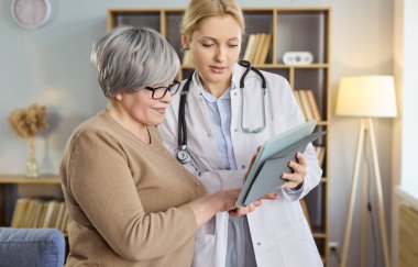 Doktor ve kıdemli hasta tablet kullanıyor. Ziyaret sırasında evde test sonuçlarını gözden geçirir ve sonraki adımları açıklar, tavsiye desteği ve bakım sunarlar. Yardımcı ev danışmanlığı kavramı.